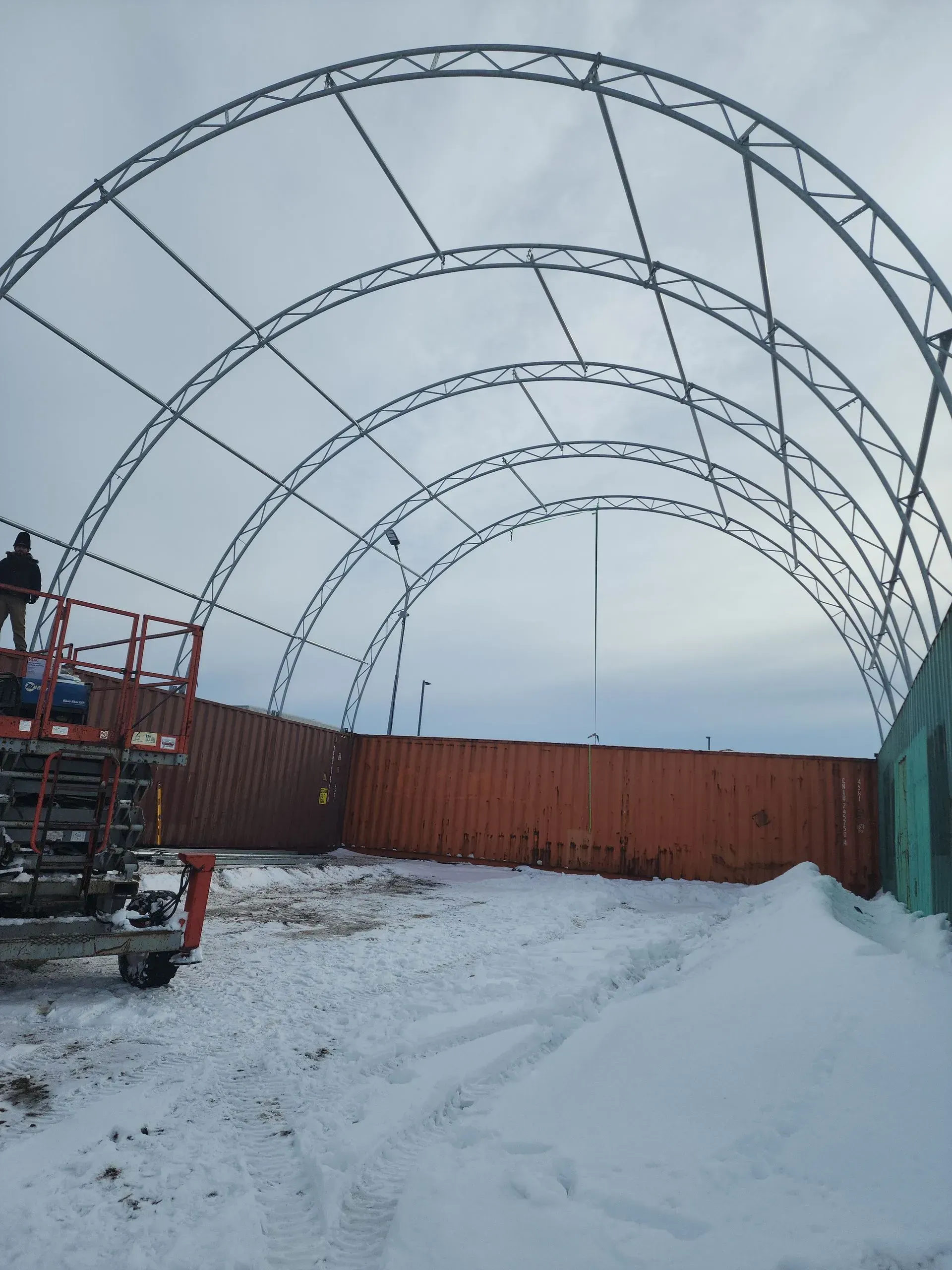 Snowy construction site with a large metal arch frame and orange shipping containers under a gray sky