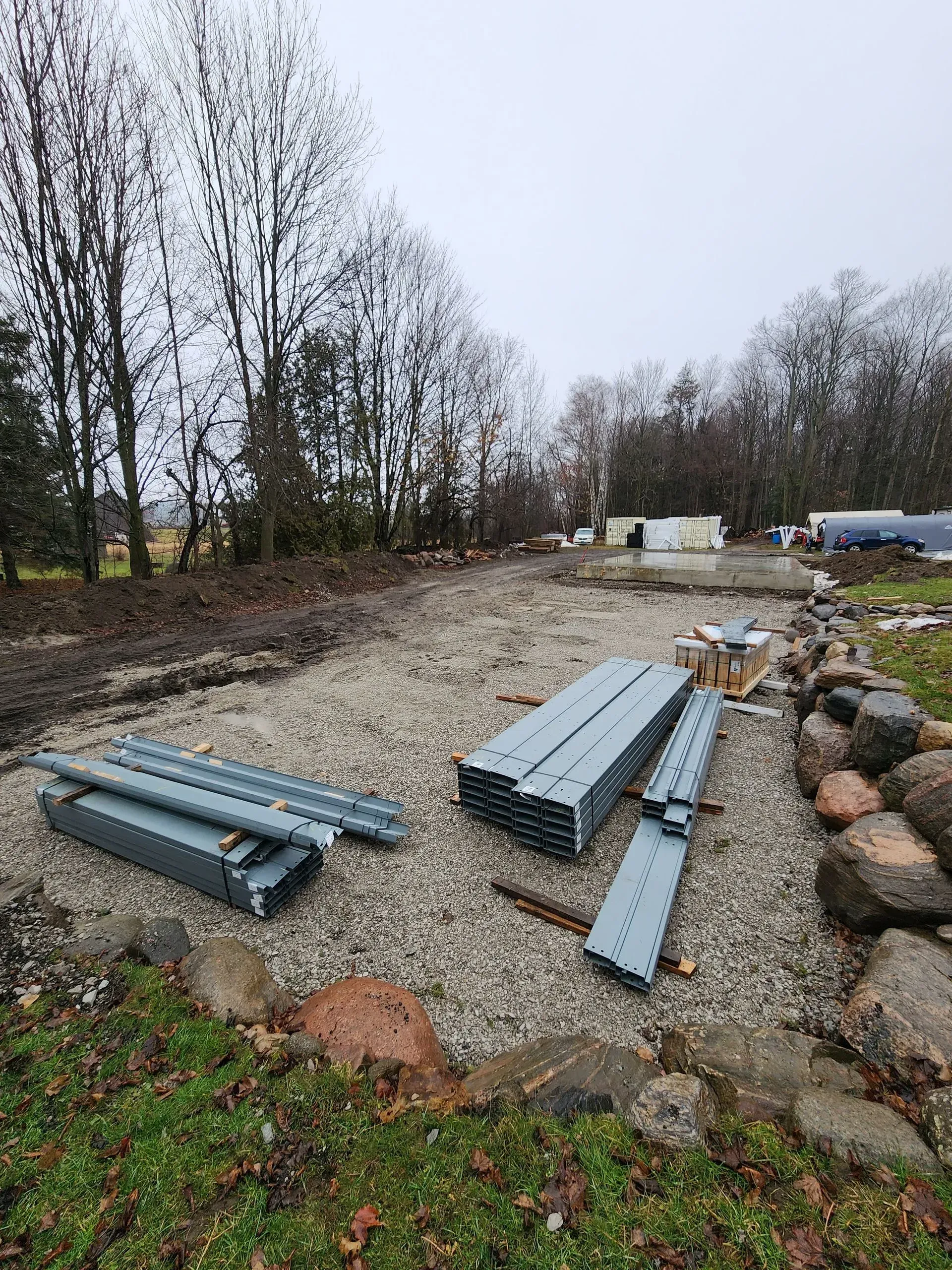 Stacks of blue-gray metal beams on gravel in a wooded outdoor worksite with rocks and equipment.