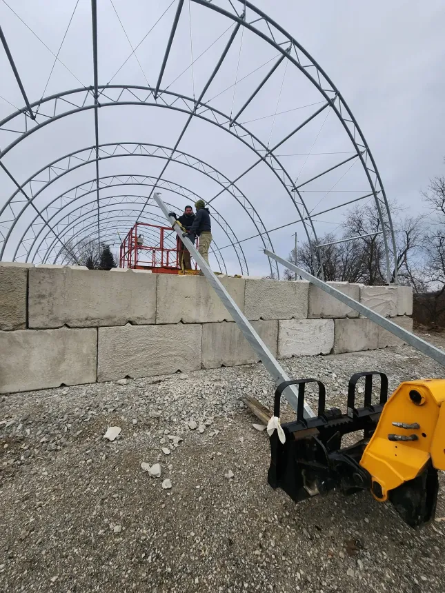 Yellow machine spraying gravel onto a stone wall beneath a large arched greenhouse frame