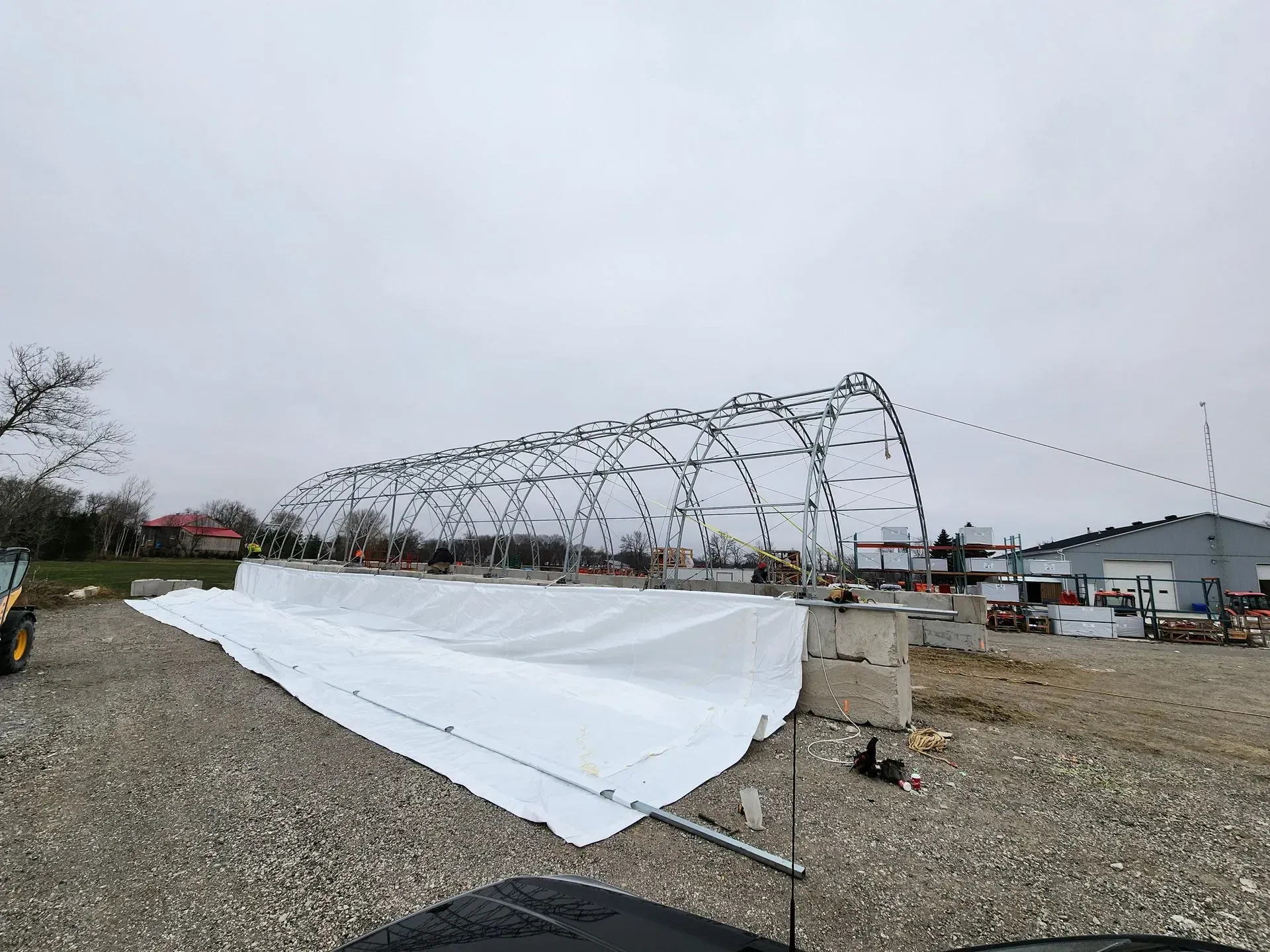 Large white tarp stretched under a metal frame in a gravel lot beside industrial buildings under an overcast sky