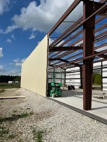 Industrial warehouse under construction with steel frame, beige wall panels, and green lift on gravel lot