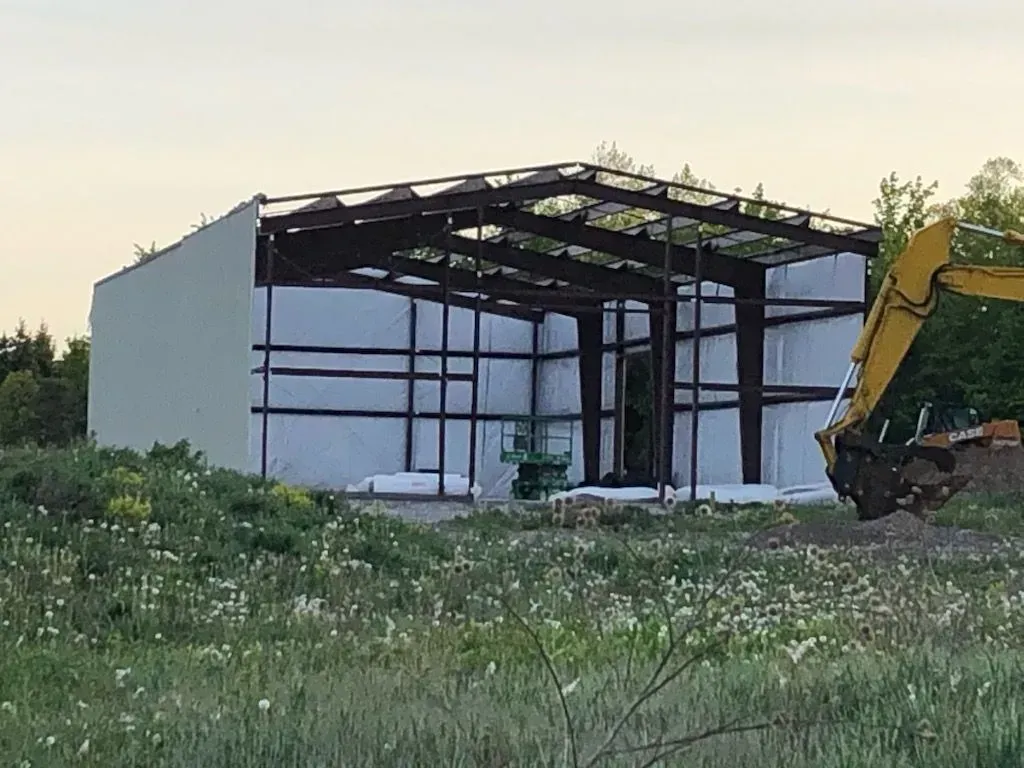 Partially built metal shed in a grassy field with excavator at right at sunset