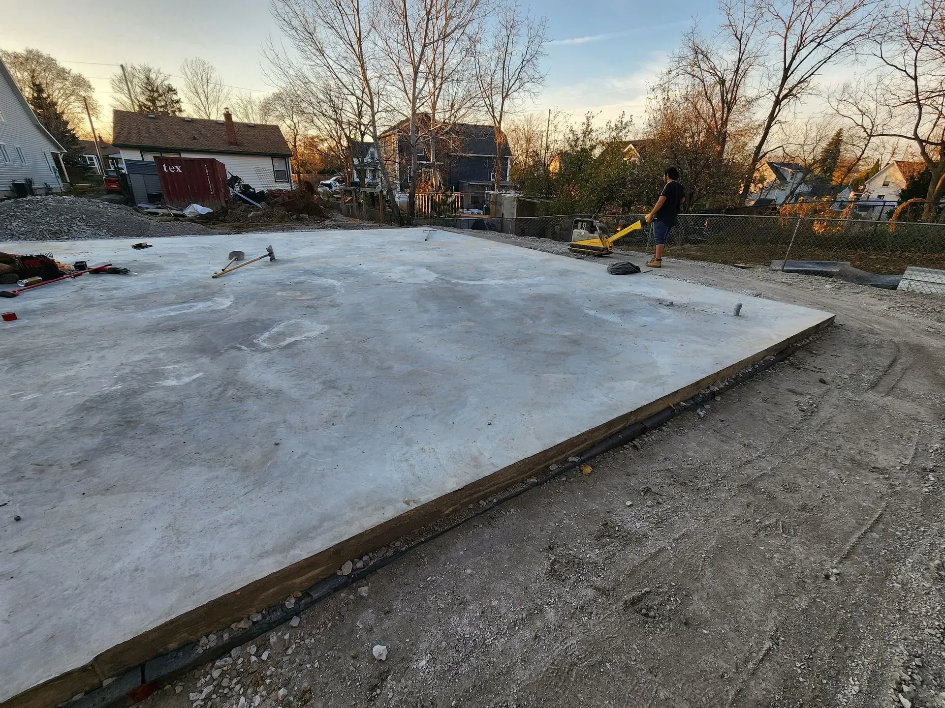 Concrete slab foundation in a dirt lot with construction equipment and bare trees at sunset