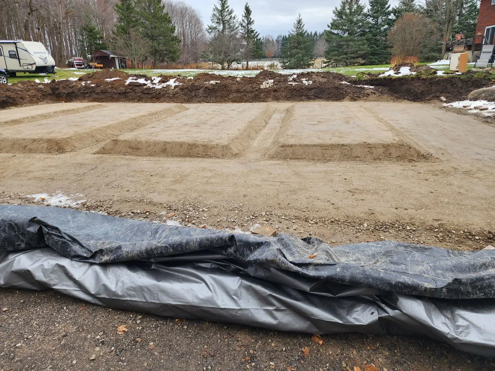 Graded construction lot with rectangular foundation forms, gravel foreground, and trees in the background