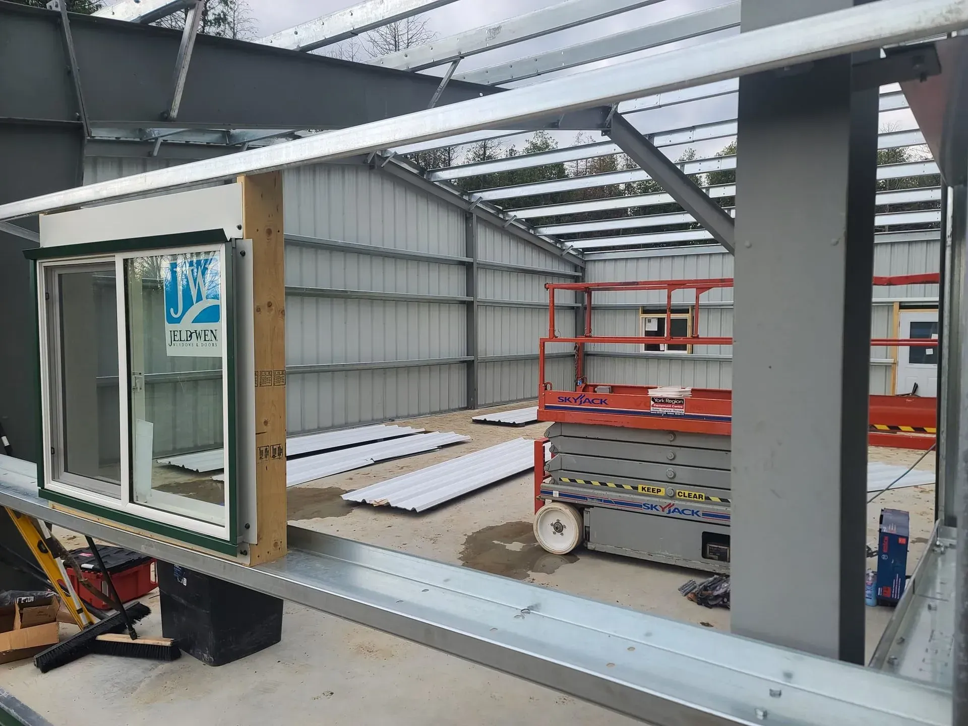 Partially built window module in a metal-framed industrial workshop with a red scissor lift in the background