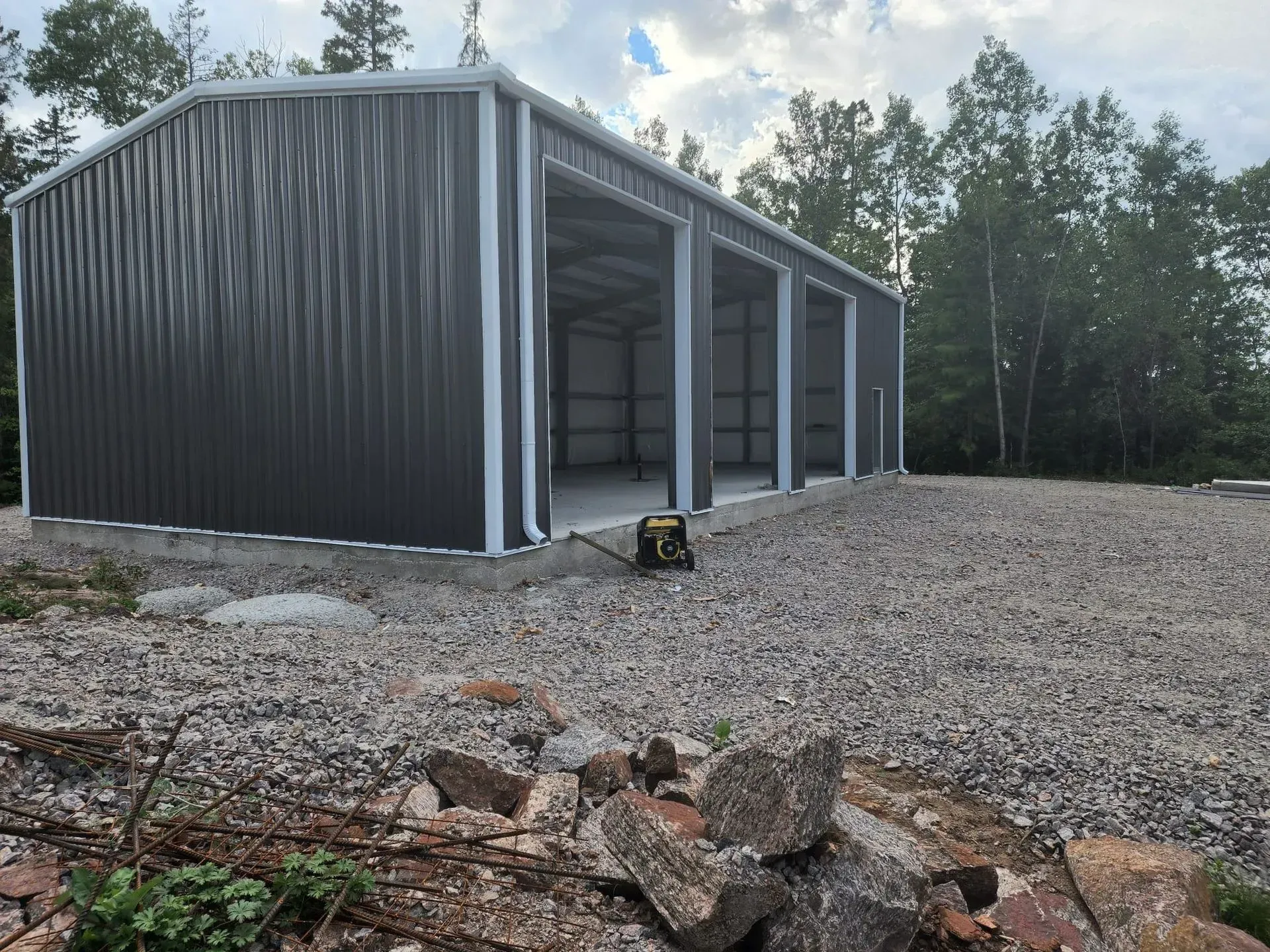 Large gray metal garage under construction on a gravel lot, with trees in the background.