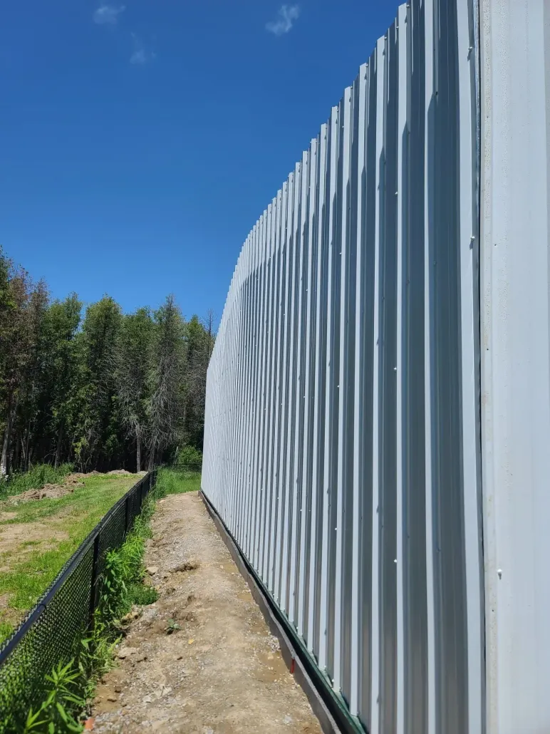 Long white corrugated fence beside a dirt path and green trees under a clear blue sky