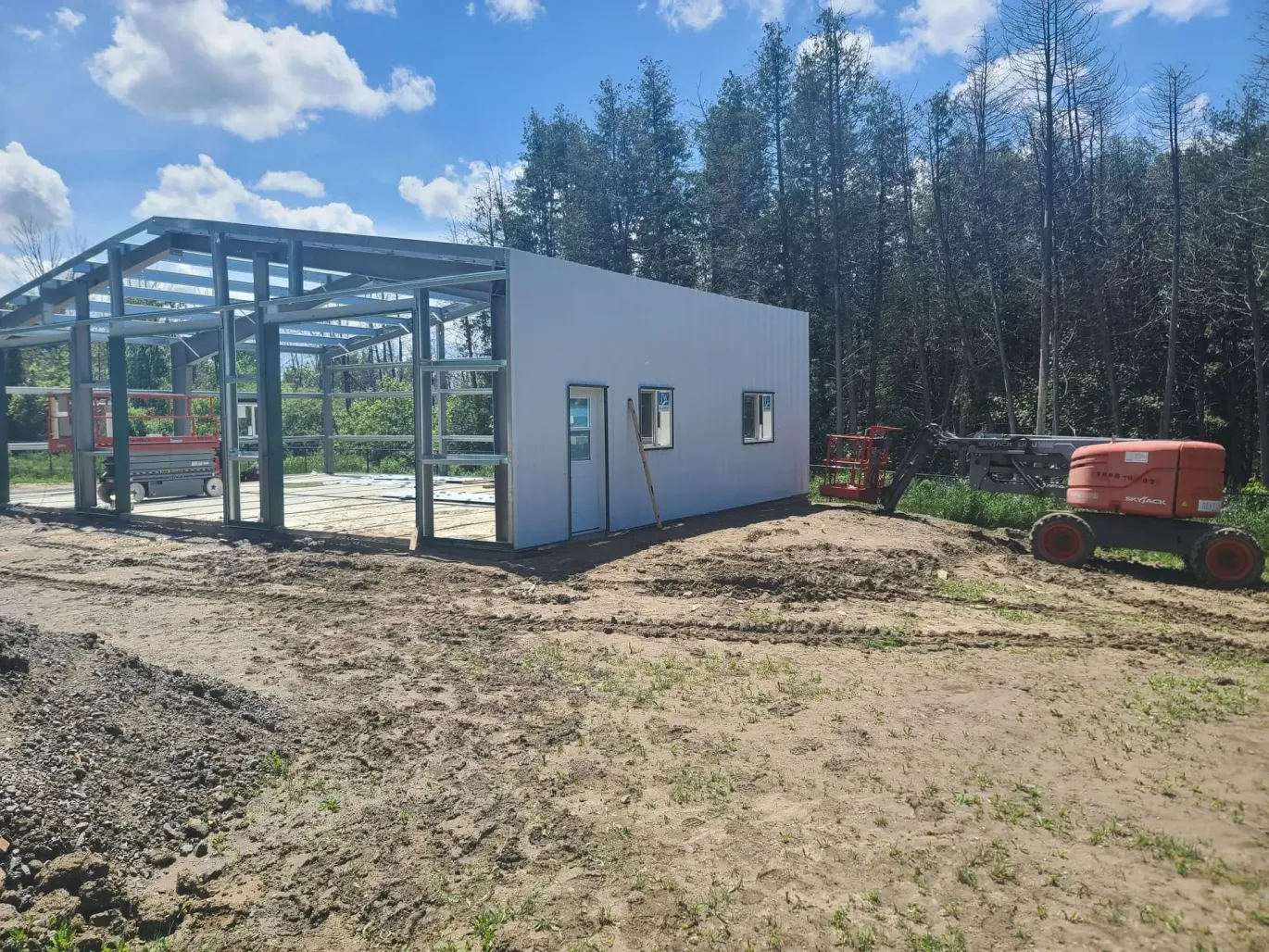 Partially built metal-framed structure beside a white modular building on a muddy construction site with a red generator.