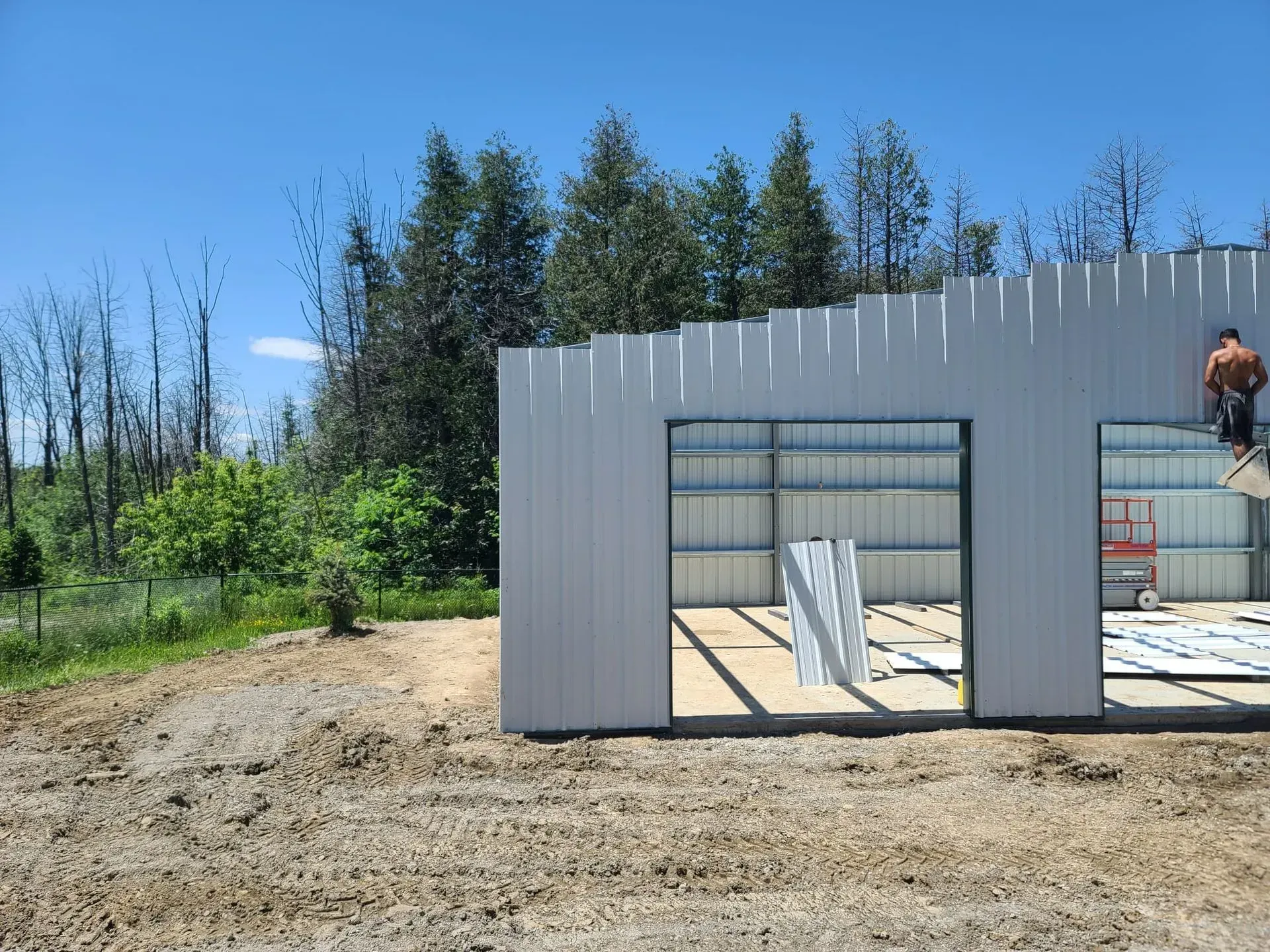 Partially built gray metal structure on a dirt lot with trees and blue sky in the background