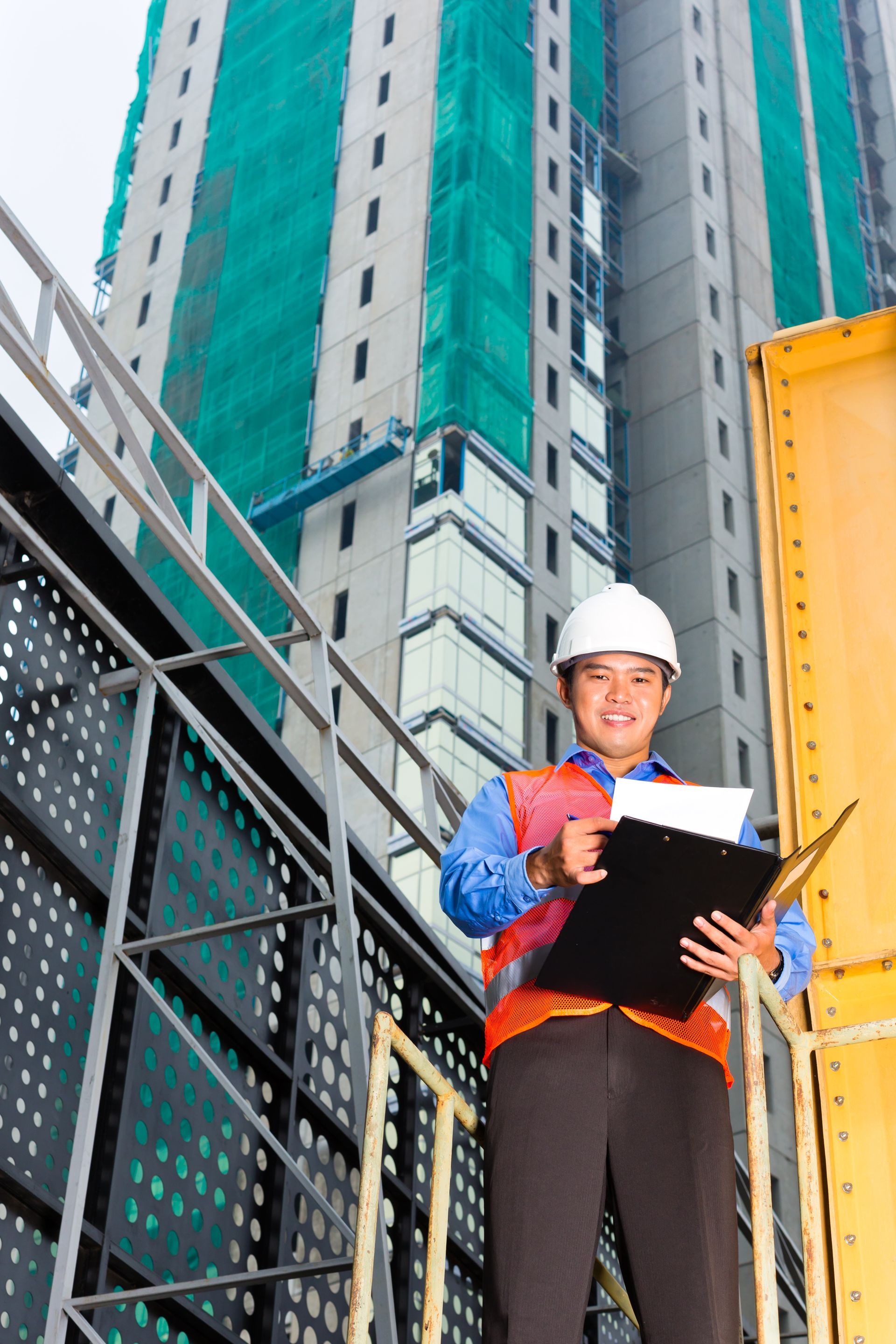Construction worker in hard hat holding clipboard beside tall building and crane