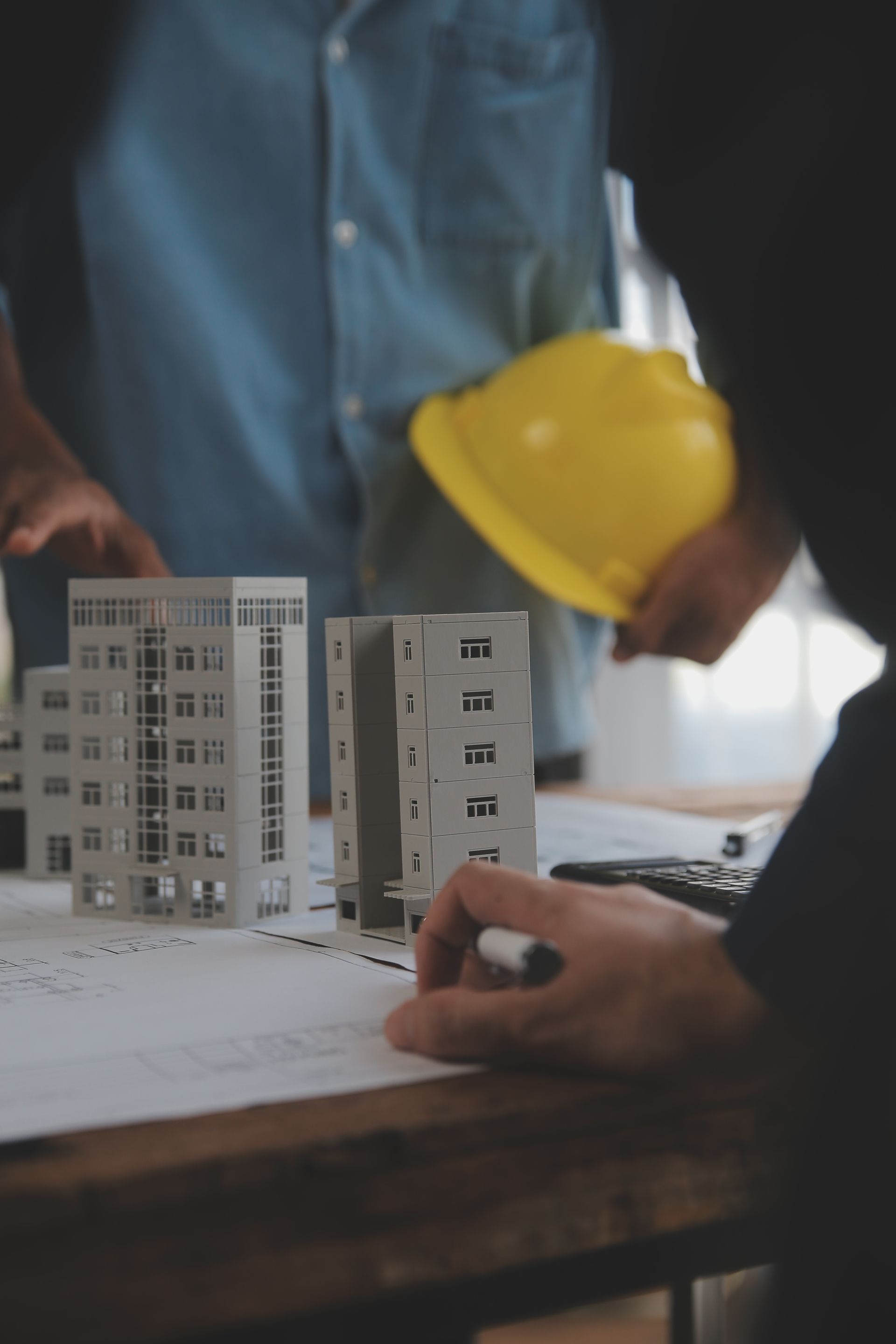 People reviewing a scale model building and blueprints at a construction meeting, with a yellow hard hat visible.