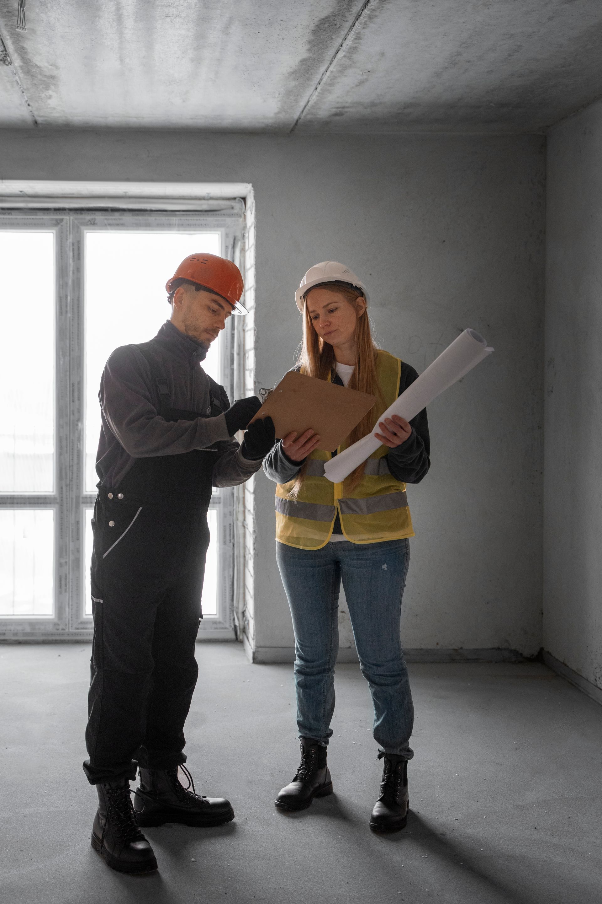 Two workers in hard hats review a blueprint inside an unfinished room.