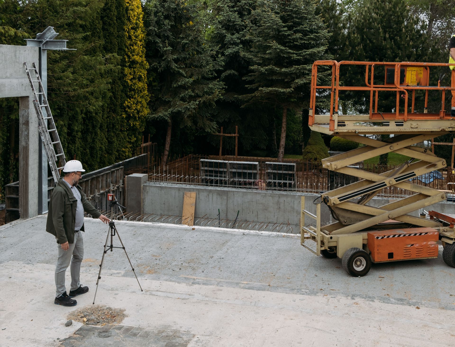 Worker standing beside a scissor lift at a construction site with a concrete foundation and trees in the background