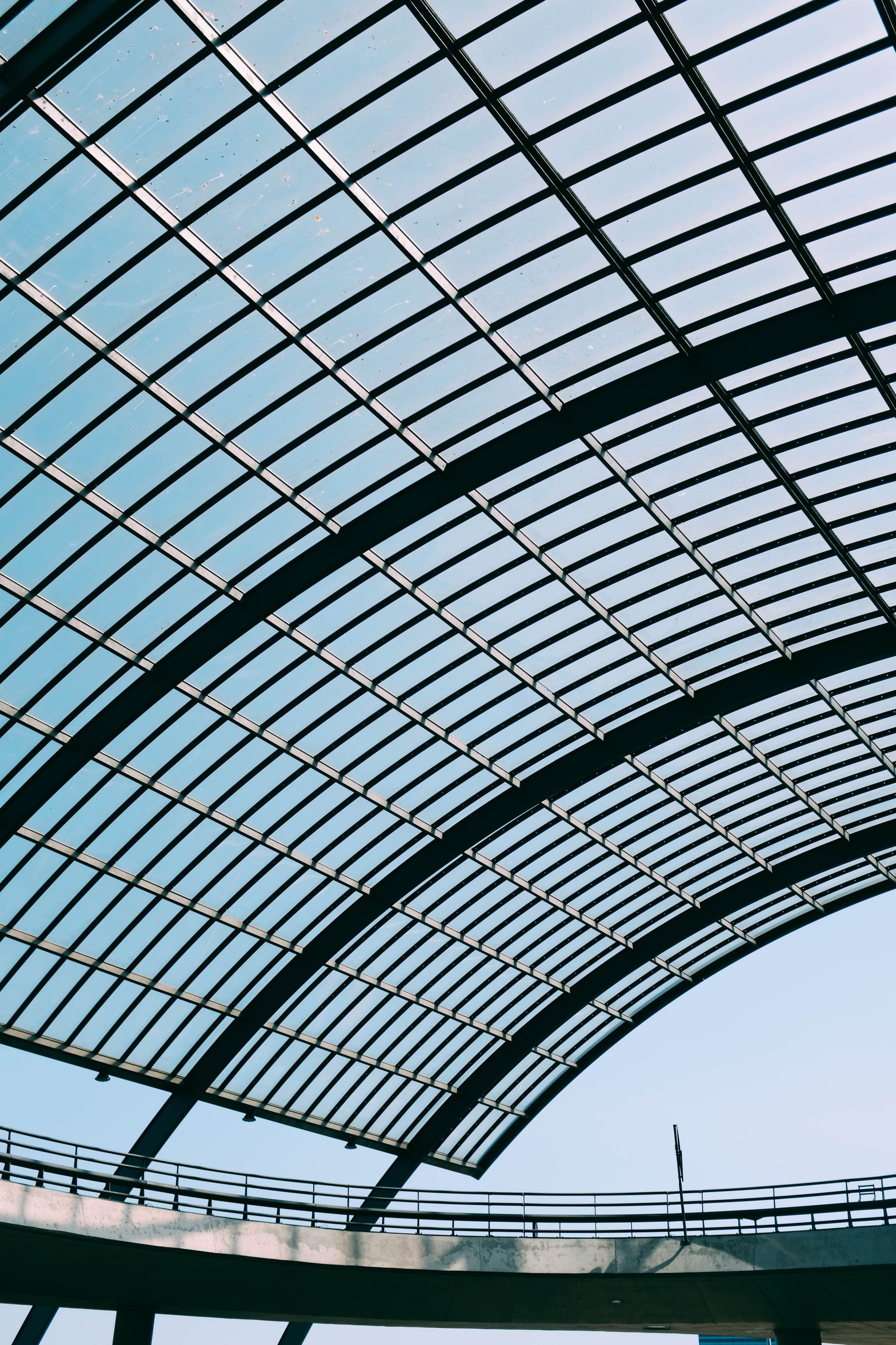 Curved glass roof canopy against a pale blue sky, viewed from below