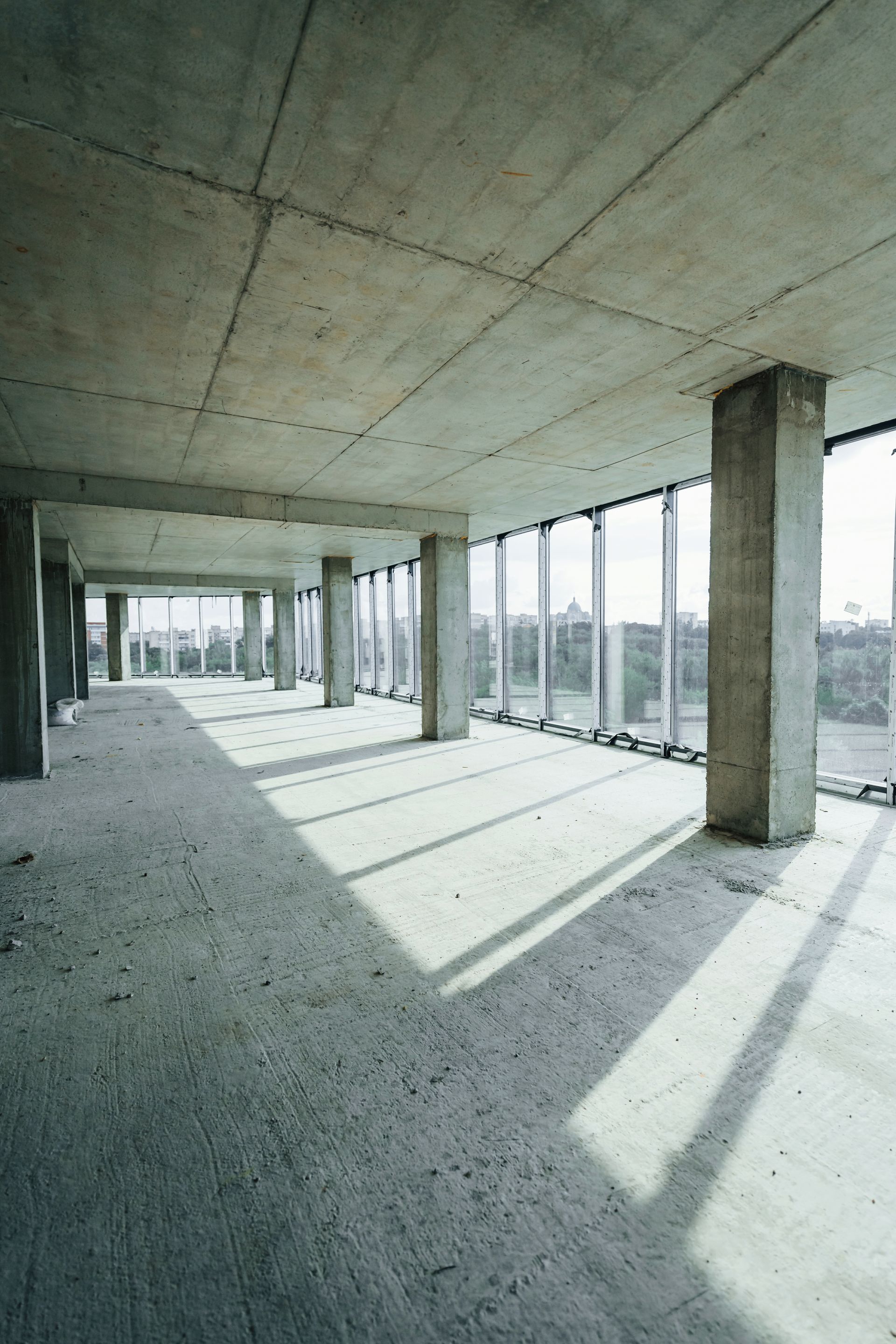 Empty concrete interior with tall columns and floor-to-ceiling windows casting long shadows.