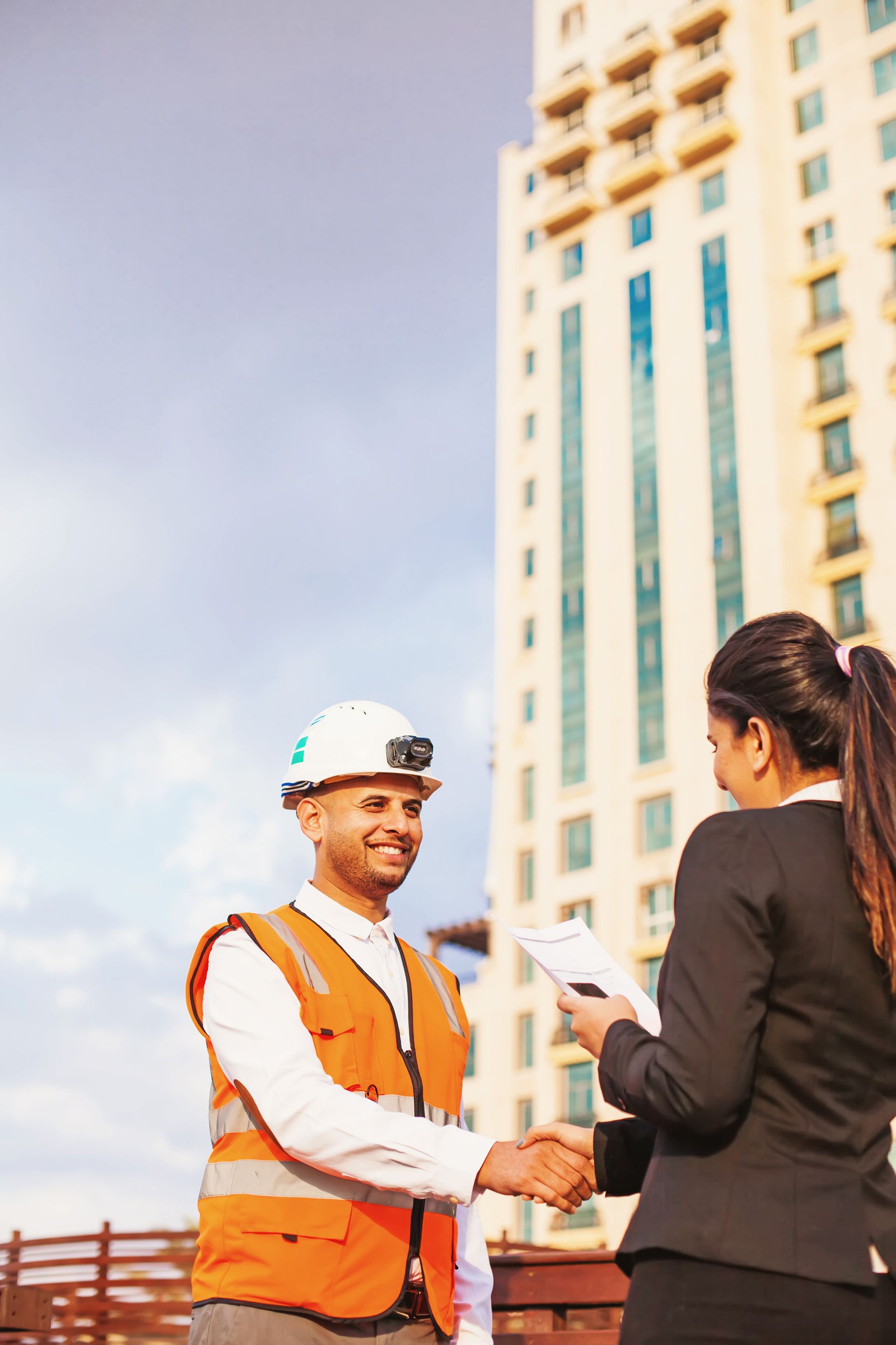 Worker in orange safety vest shaking hands with a businesswoman outside a high-rise building