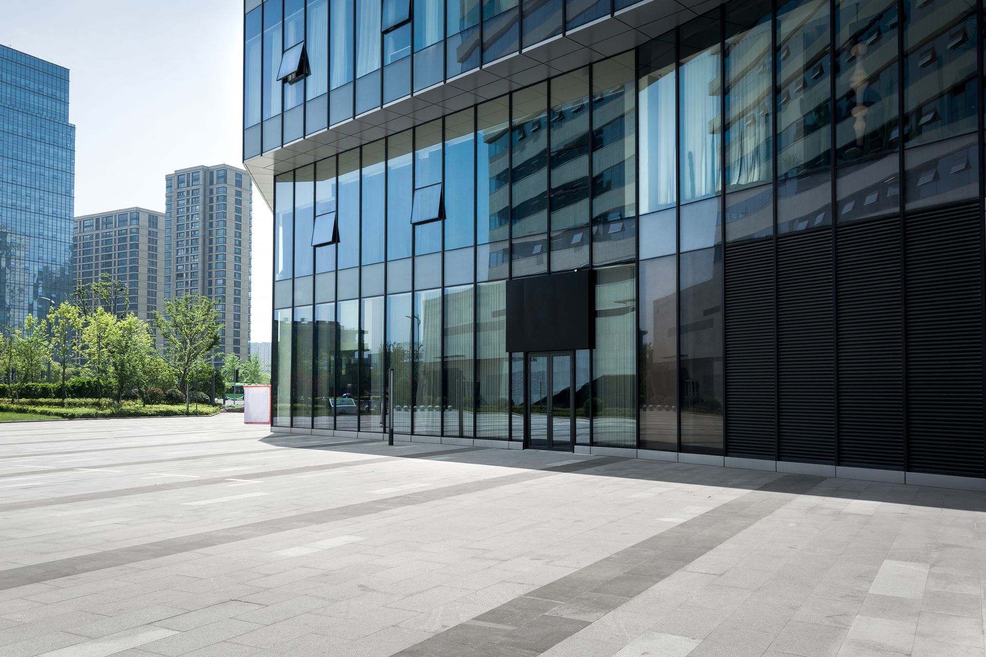 Modern glass office building beside a sunlit plaza with trees and high-rises in the background