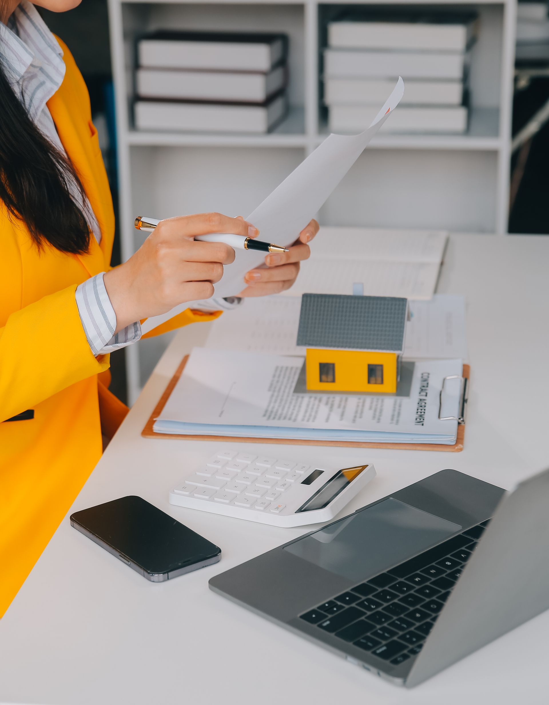 Person in yellow jacket reviewing papers at desk with laptop and small house model