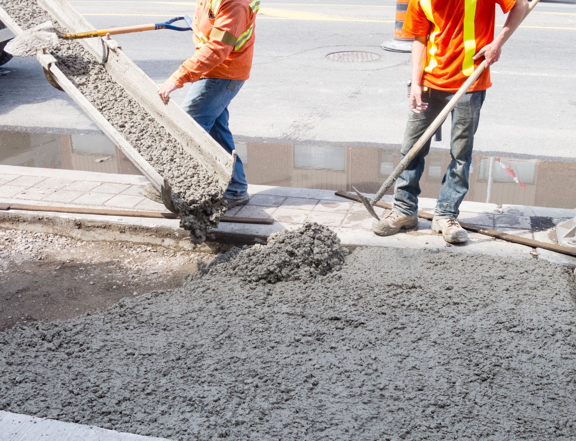 Workers in orange vests pour wet concrete at a road construction site.