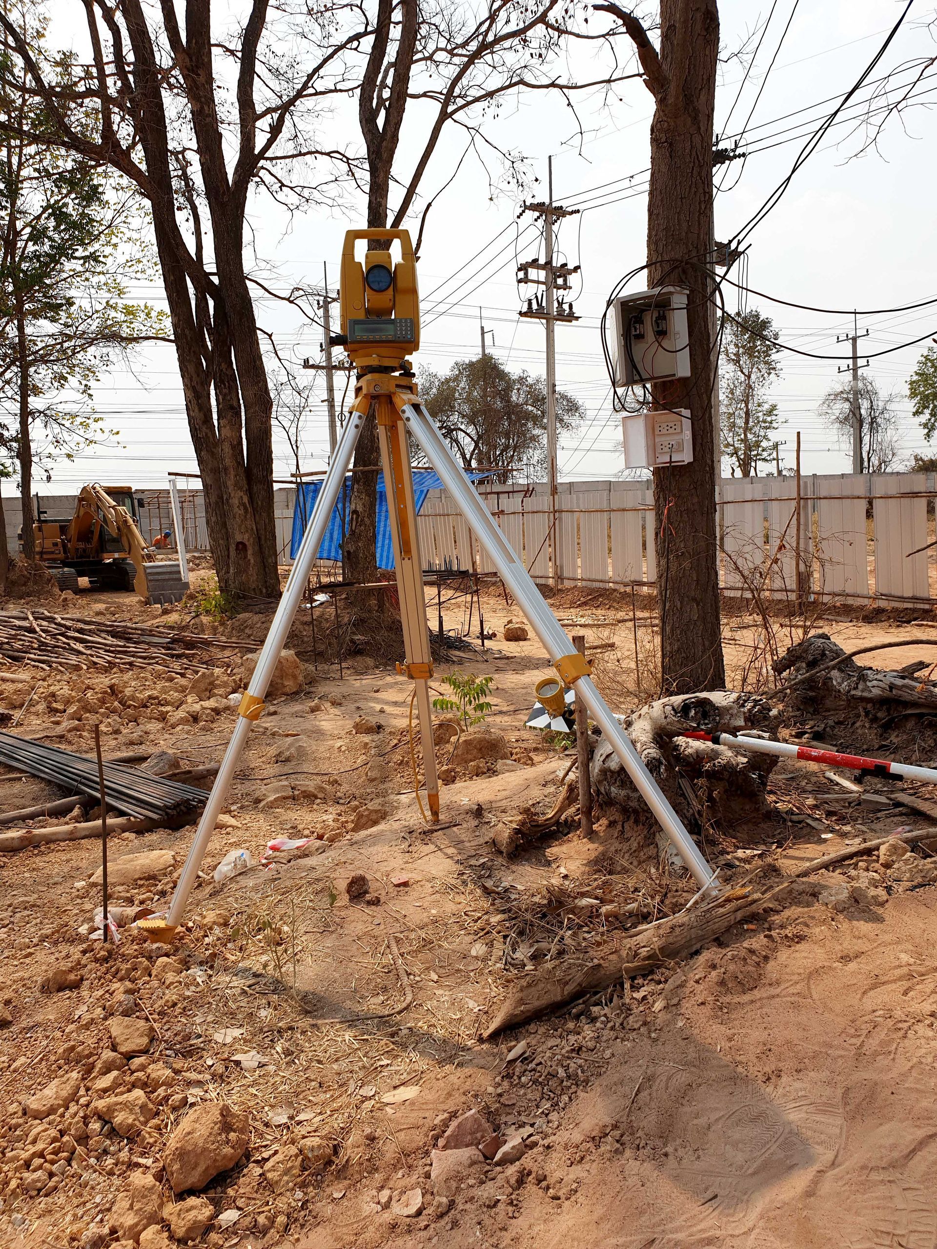 Surveying tripod with mounted instrument on a muddy construction site near trees and utility poles