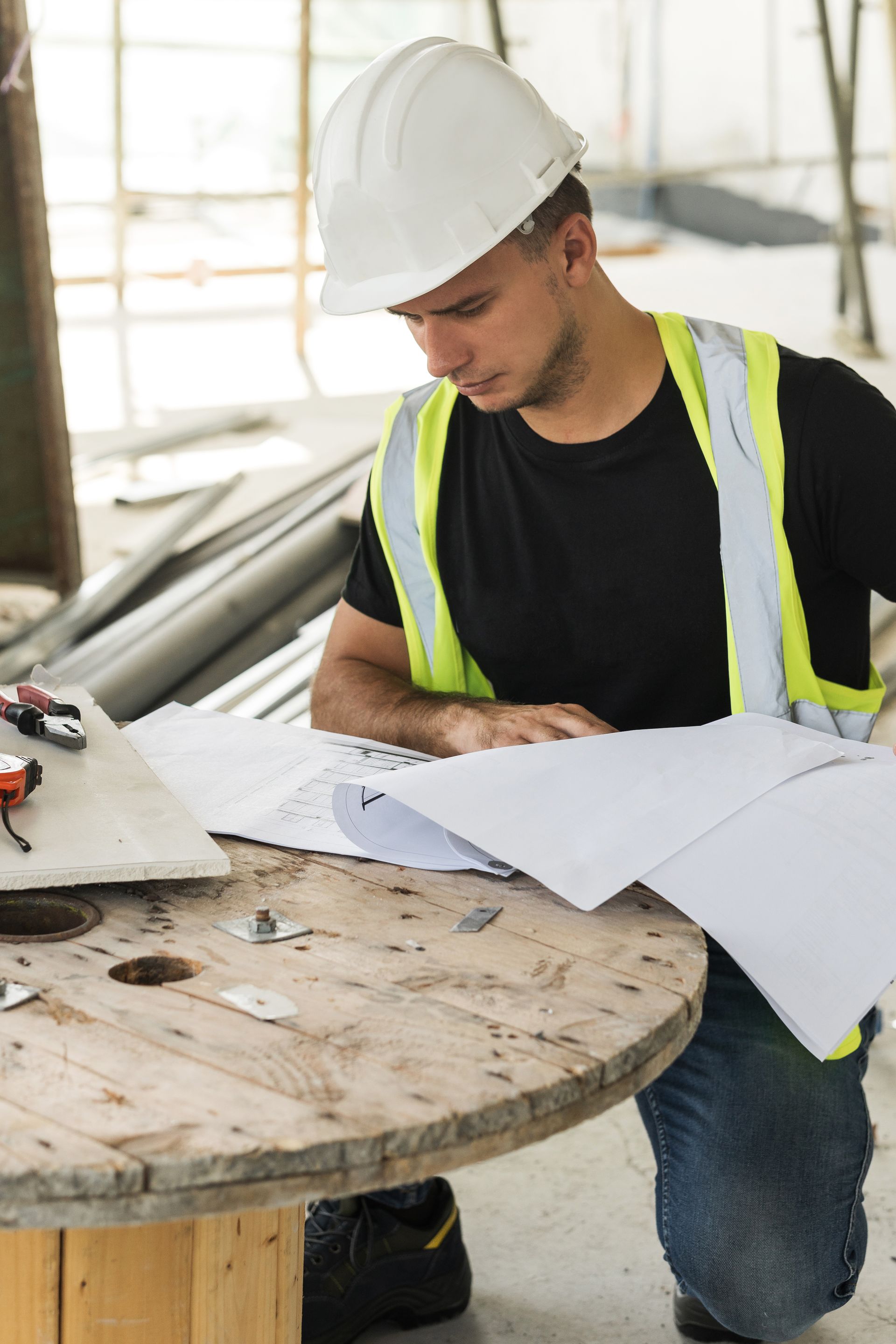 Construction worker in hard hat and safety vest reviewing blueprints at a worksite table
