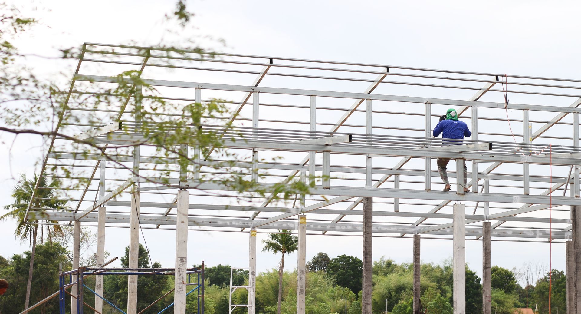 Worker on a white steel frame roof, standing on beams amid greenery under construction.