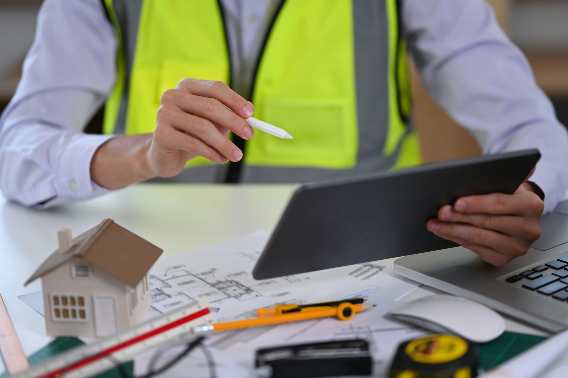 Person in a safety vest reviewing a house model and blueprint with a laptop and measuring tools on a desk