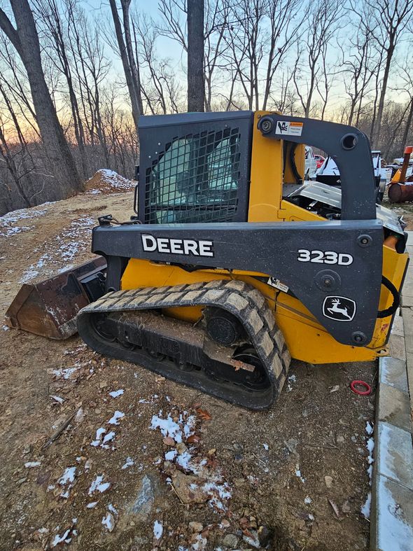 John Deere 323D compact track loader, yellow and black, parked outdoors on dirt with trees in the background.