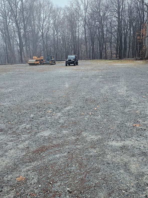 Gravel lot with vehicles: a black SUV, an excavator, and a motorcycle, with a blurred forest background.