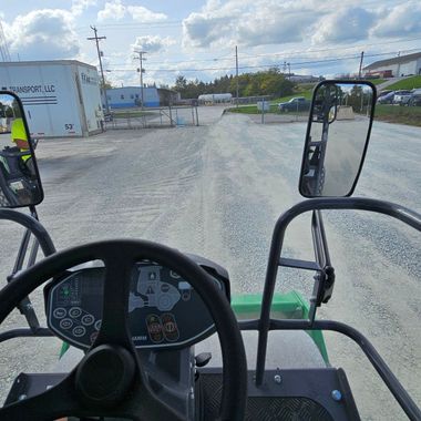 View from inside a machine driving on a gravel road toward a gate, with a worker visible on the left side.