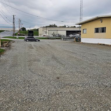 Gravel parking area next to a building and road, overcast sky. A dark car is in the parking area.