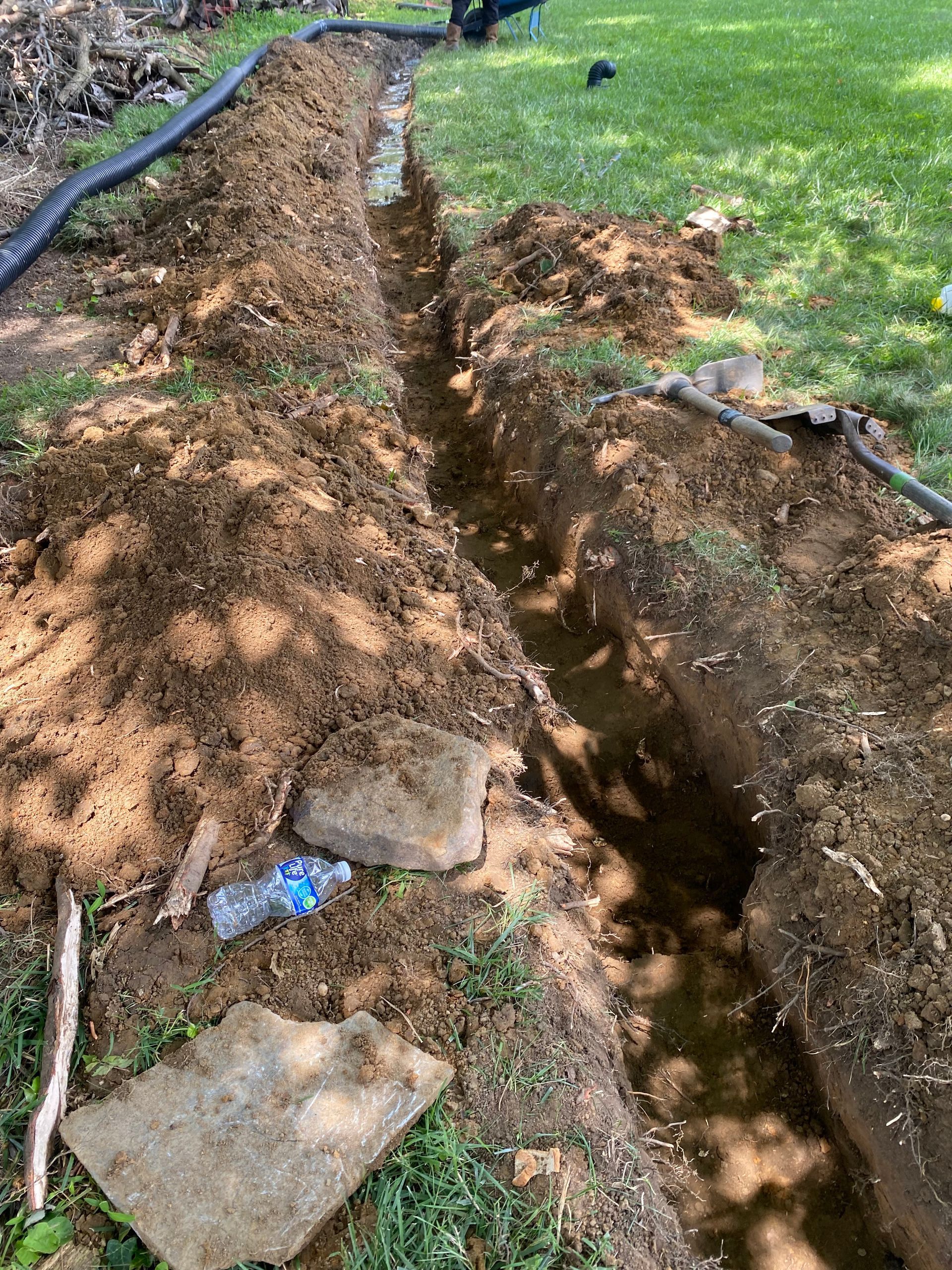 Trench dug in a grassy yard, filled with water, near a black hose. Dirt piles surround it
