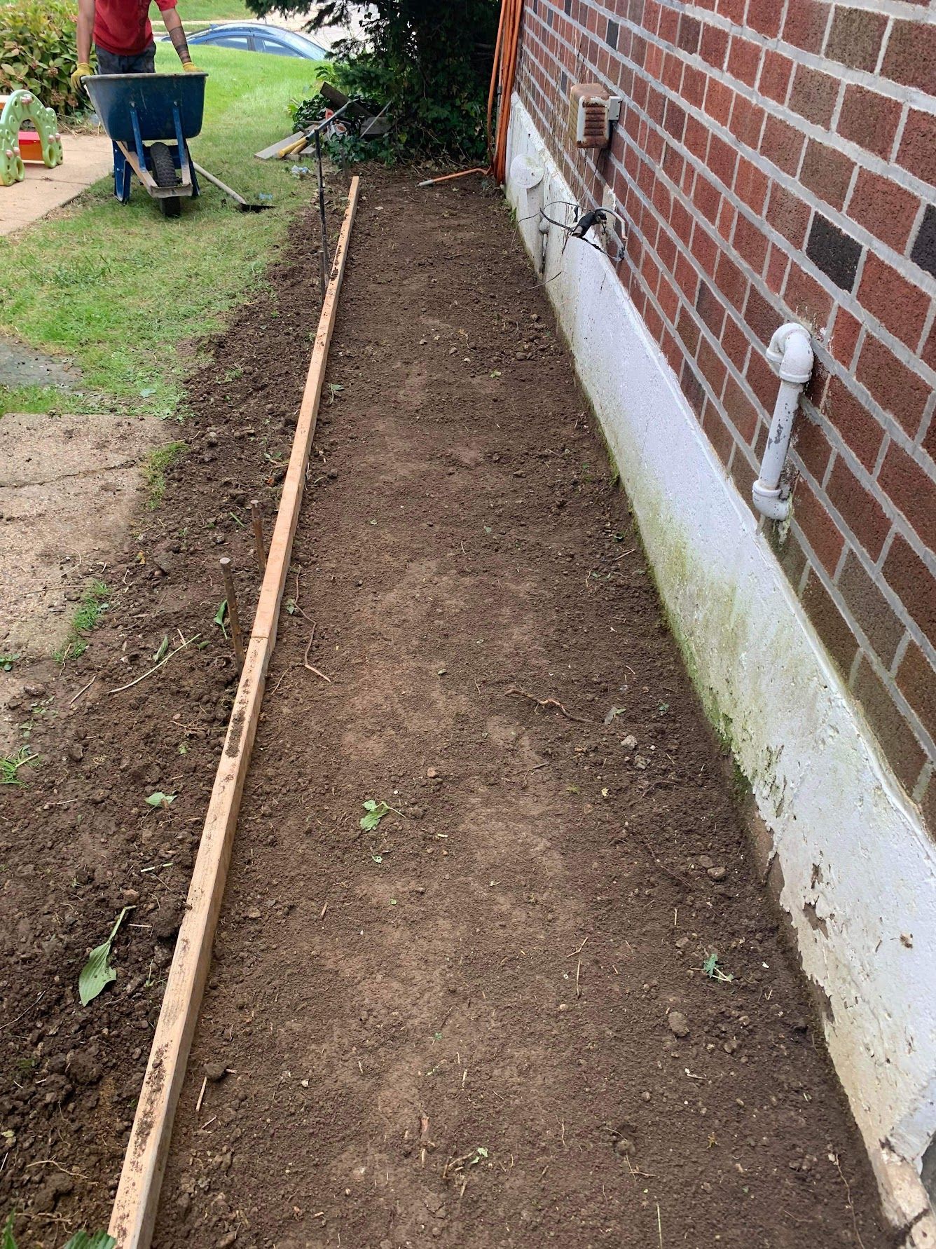 Dirt path next to brick building, wooden border on one side, person using a wheelbarrow in background