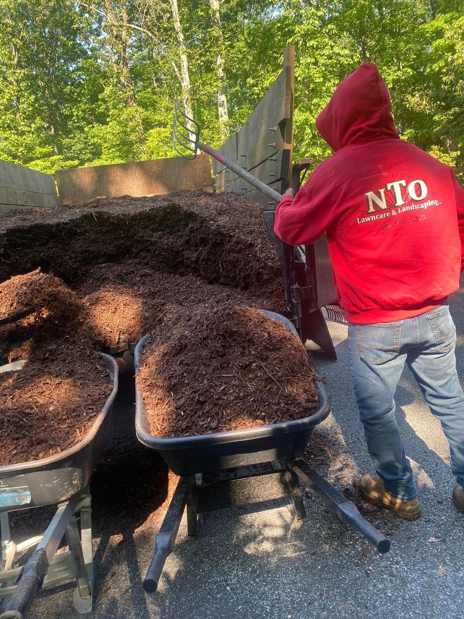 An employee wearing a red nto lawn care & landscaping hoodie is pushing a wheelbarrow full of mulch