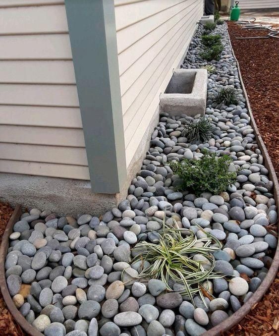 A landscaped garden bed with grey river rocks, plants, and a light tan house siding