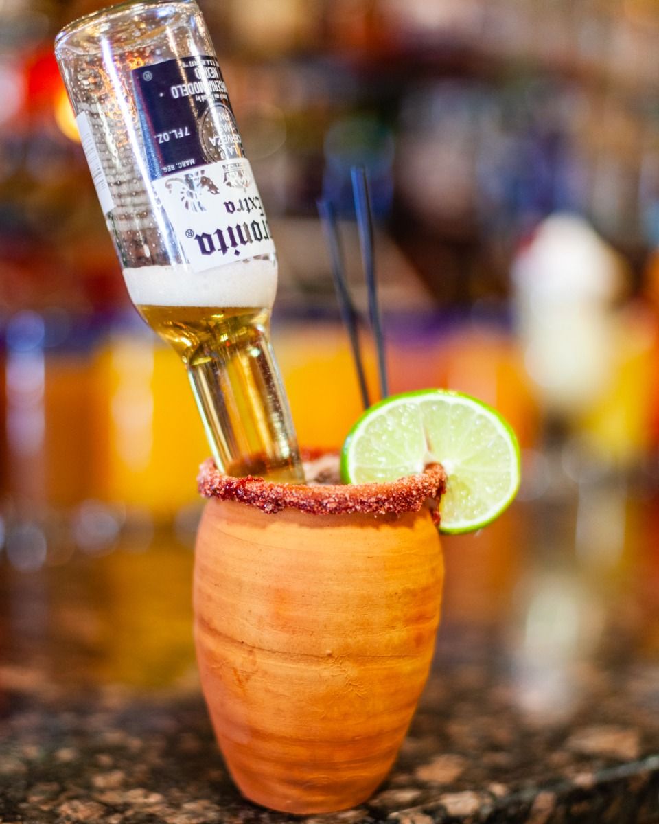 A clay mug with a beer bottle inserted, lime slice, and straws; rim has a red spice.