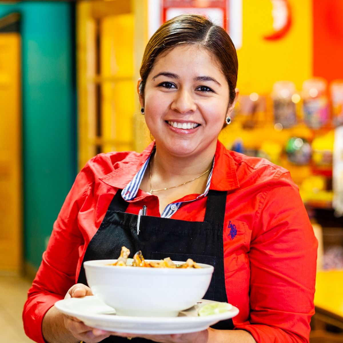 Woman holding bowl of food, smiling in a restaurant setting, wearing red shirt and black apron.