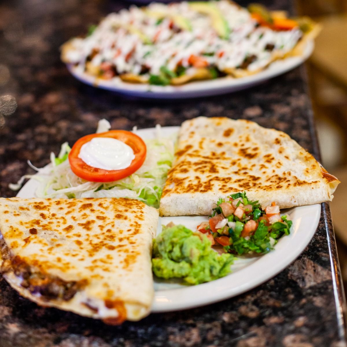 Quesadillas with guacamole and pico de gallo on a plate, with a plate of nachos in the background.