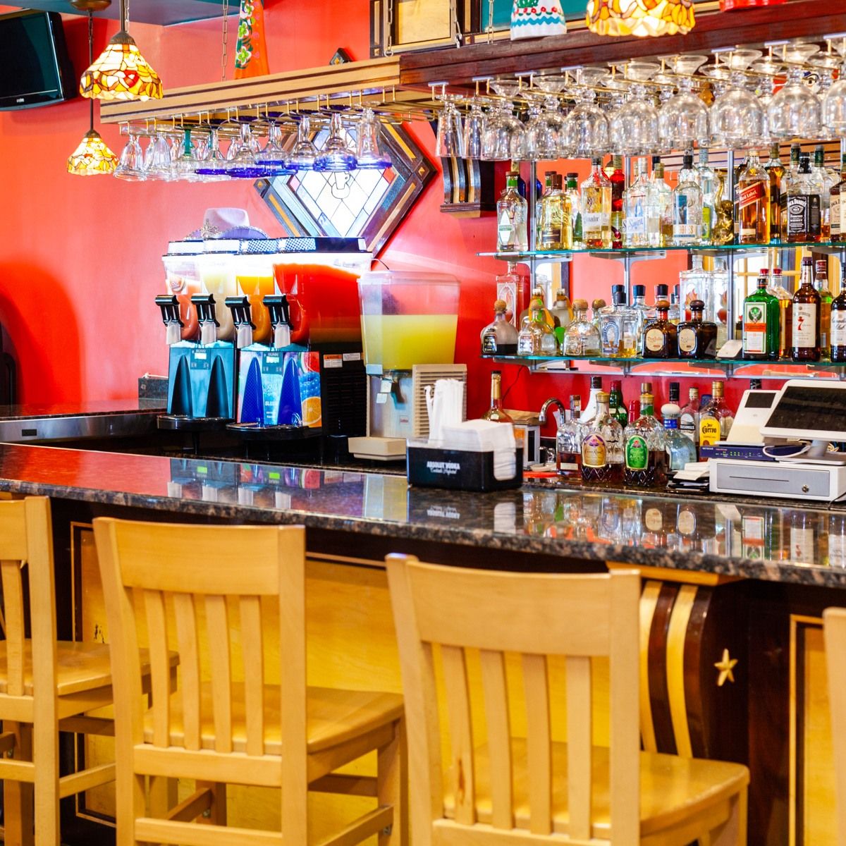 Bar with wooden stools, shelves of alcohol, and frozen drink dispensers.