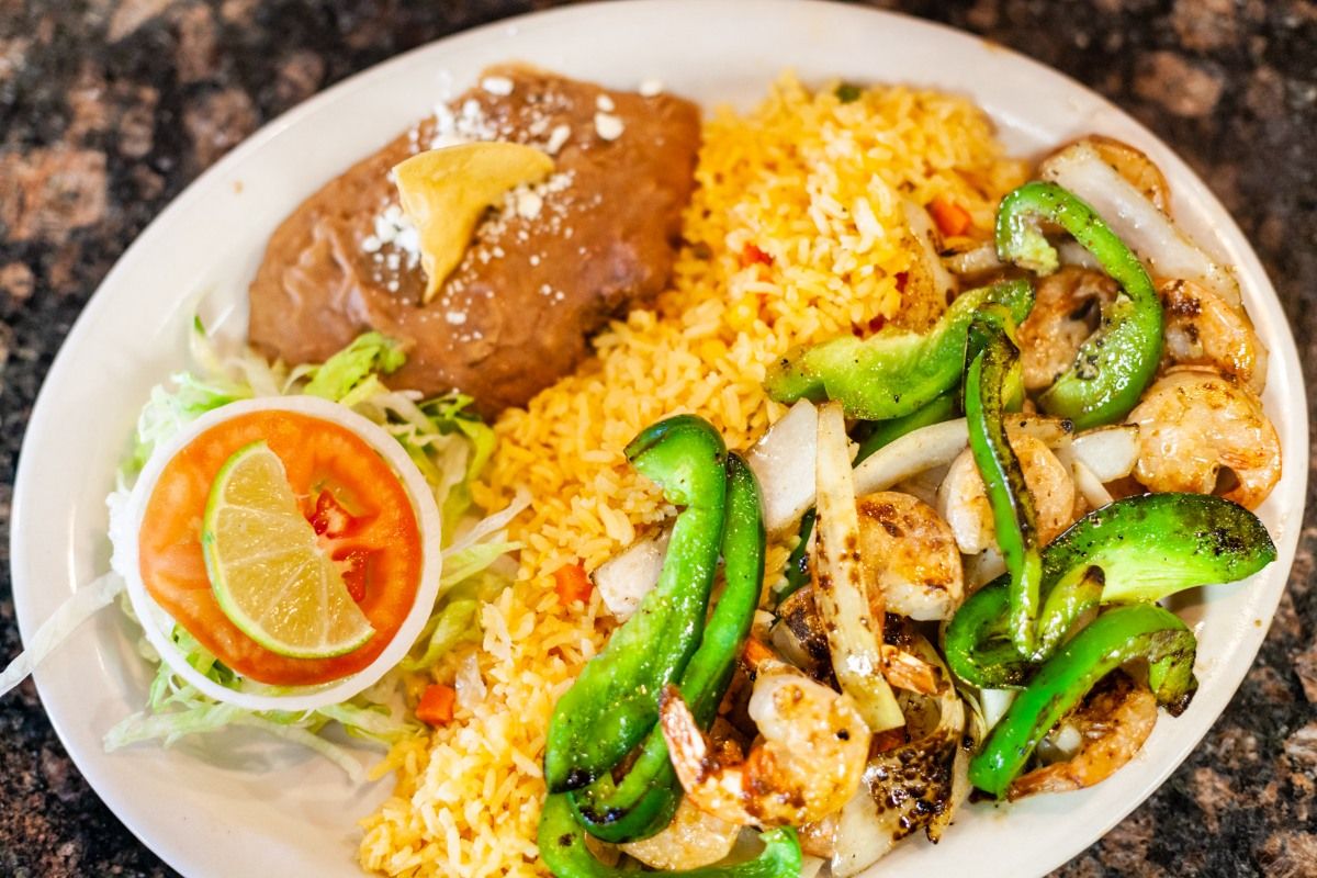 Plate with Mexican food: refried beans, rice, grilled shrimp and peppers, lettuce, tomato, lime.