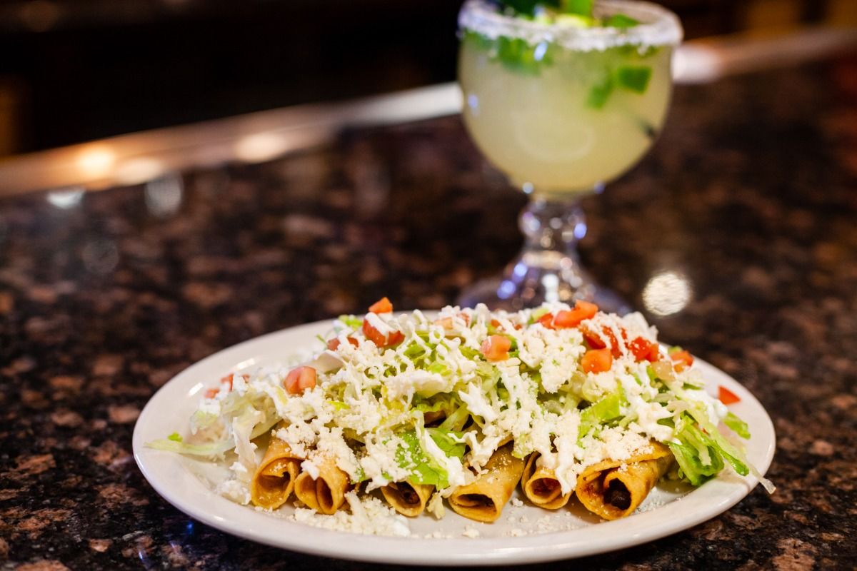 Plate of rolled tacos topped with lettuce, tomatoes, and cheese, with a margarita in the background.