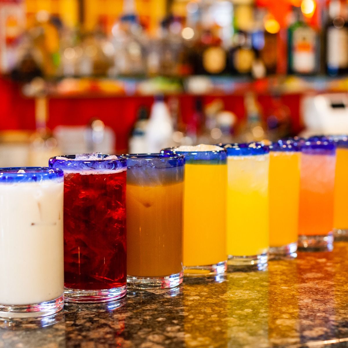 Row of colorful drinks on a bar with a blurred background of bottles and a red shelf.