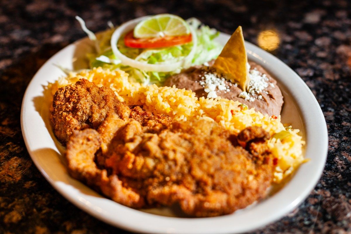 Plate of Mexican food: fried meat, yellow rice, beans with tortilla chip, lettuce, lime, and onion.