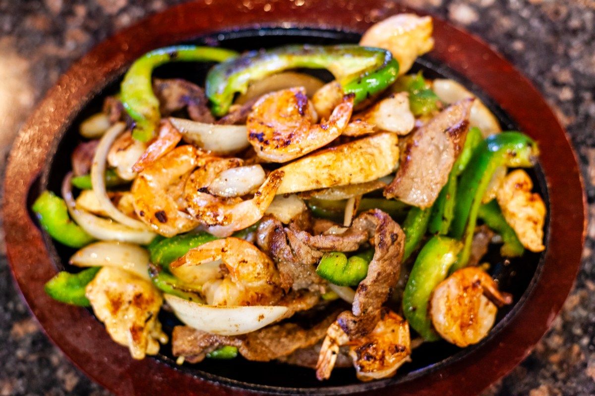 Sizzling platter of shrimp, steak, onions, and bell peppers; brown and green, served on a wooden plate.