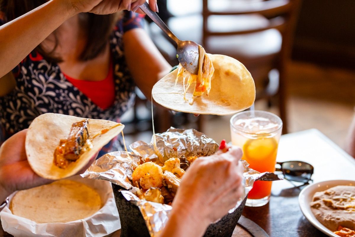 People scooping cheesy food from a bowl to fill tortillas at a restaurant table.