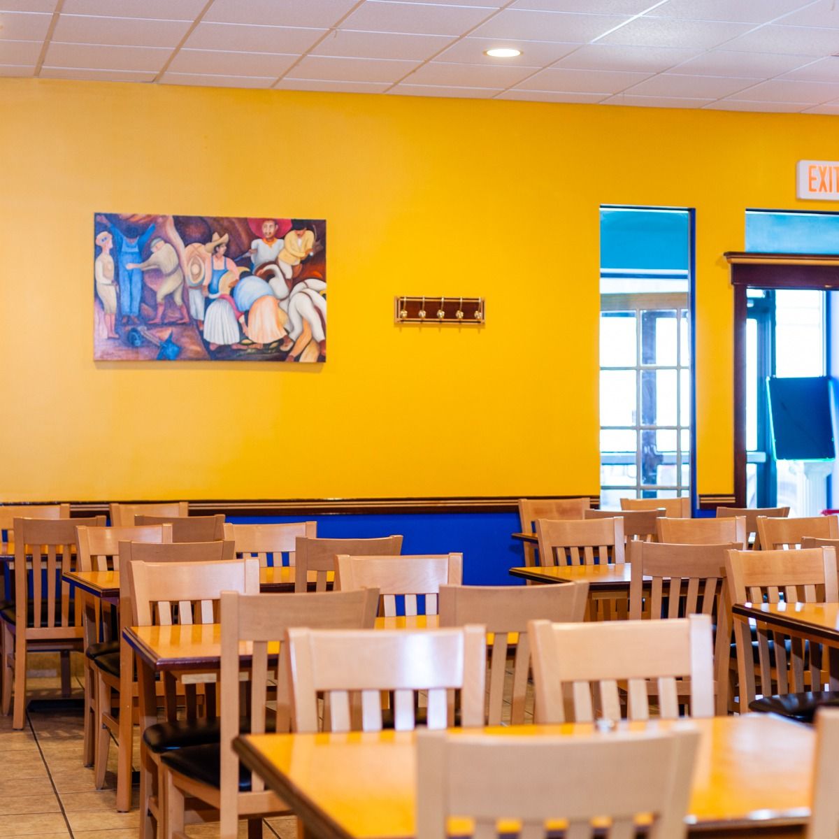 Restaurant interior with rows of wooden tables and chairs, yellow walls, and a colorful painting.