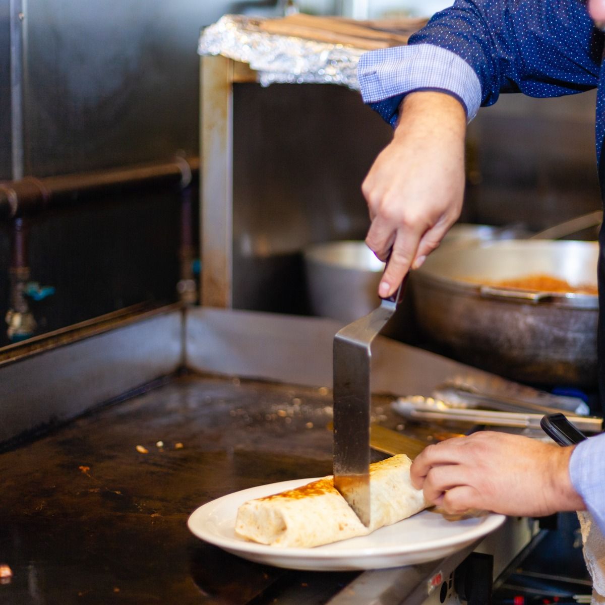 Person cutting a burrito on a plate with a spatula on a hot griddle.