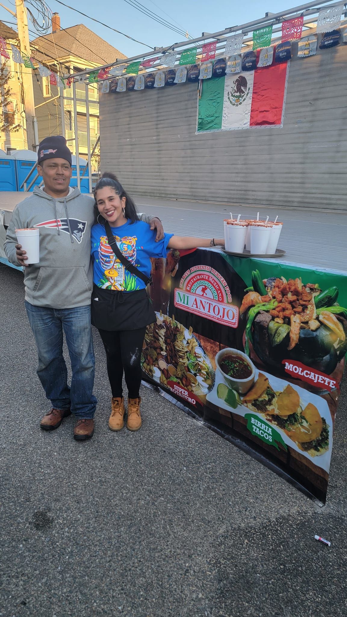 Two people pose with a taco stand sign; Mexican flag in the background.