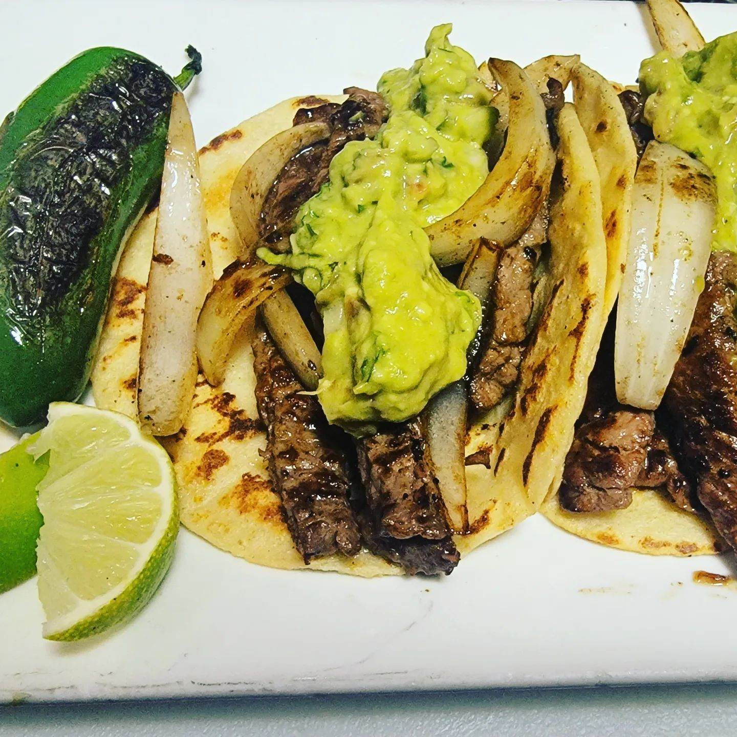 Grilled steak tacos with guacamole, onions, jalapeño, and lime slices on a white plate.