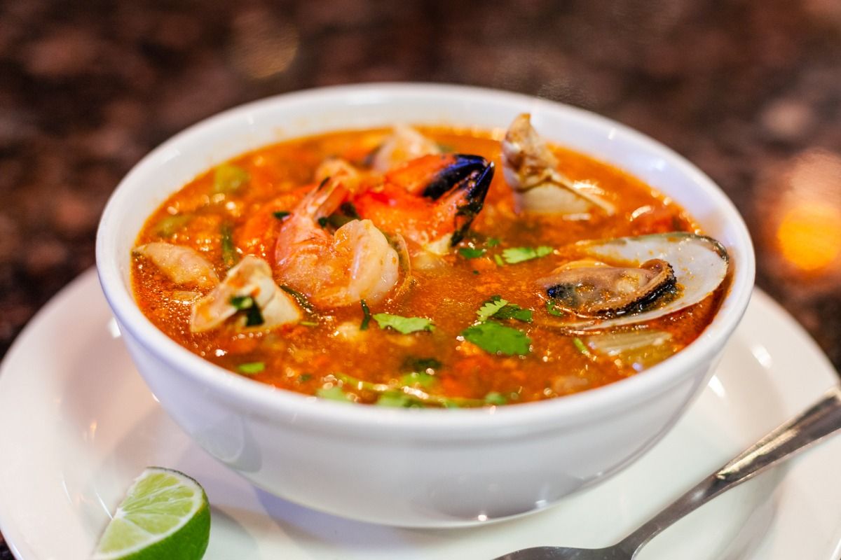 Bowl of orange seafood soup with shrimp, mussels, and lime wedge.